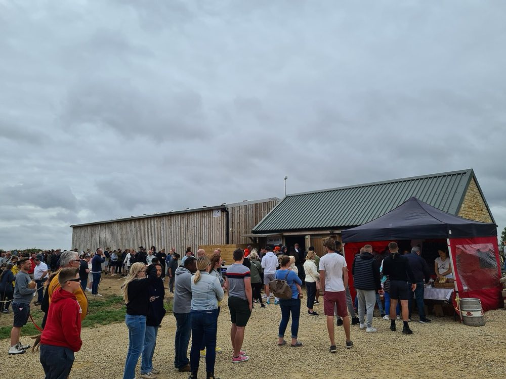 A long queue of visitors on gravel outside the green corrugated barn at Diddly Squat farm shop