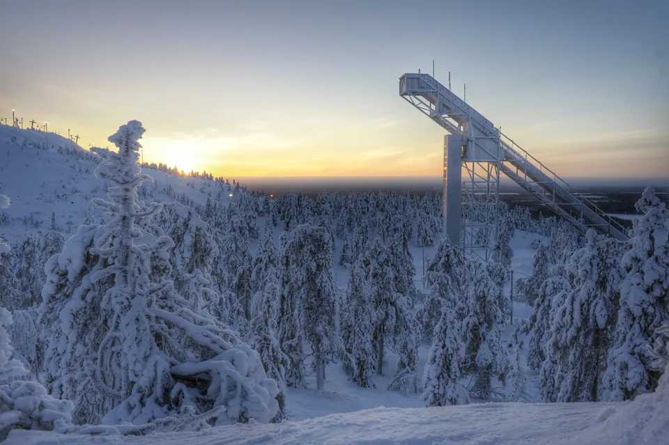 The Ruka ski jump tower above frozen forest, low sun burning gold along the horizon.